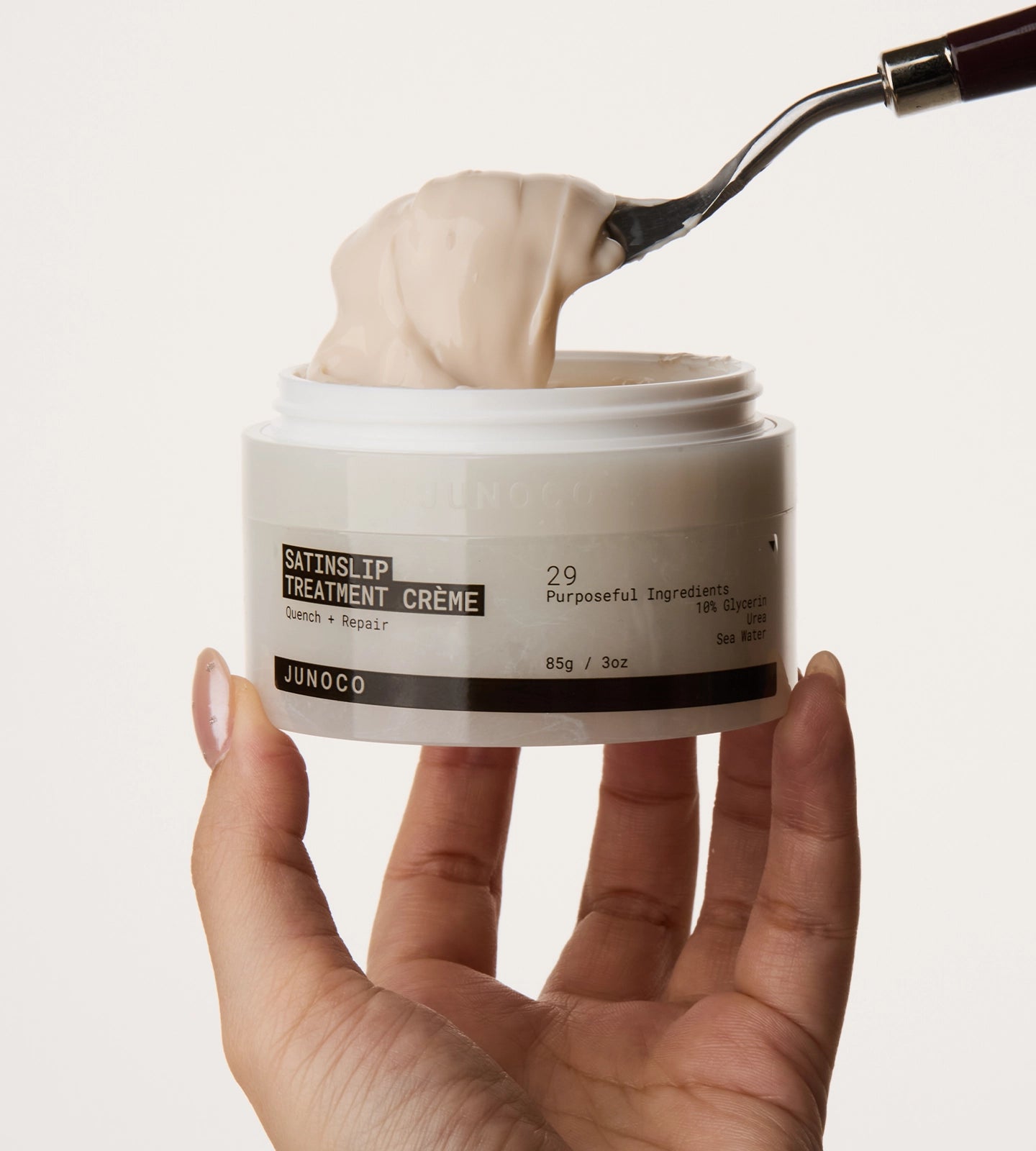 Hand holding a jar of Junoco Satin Slip Treatment Crème with a spoonful being lifted.